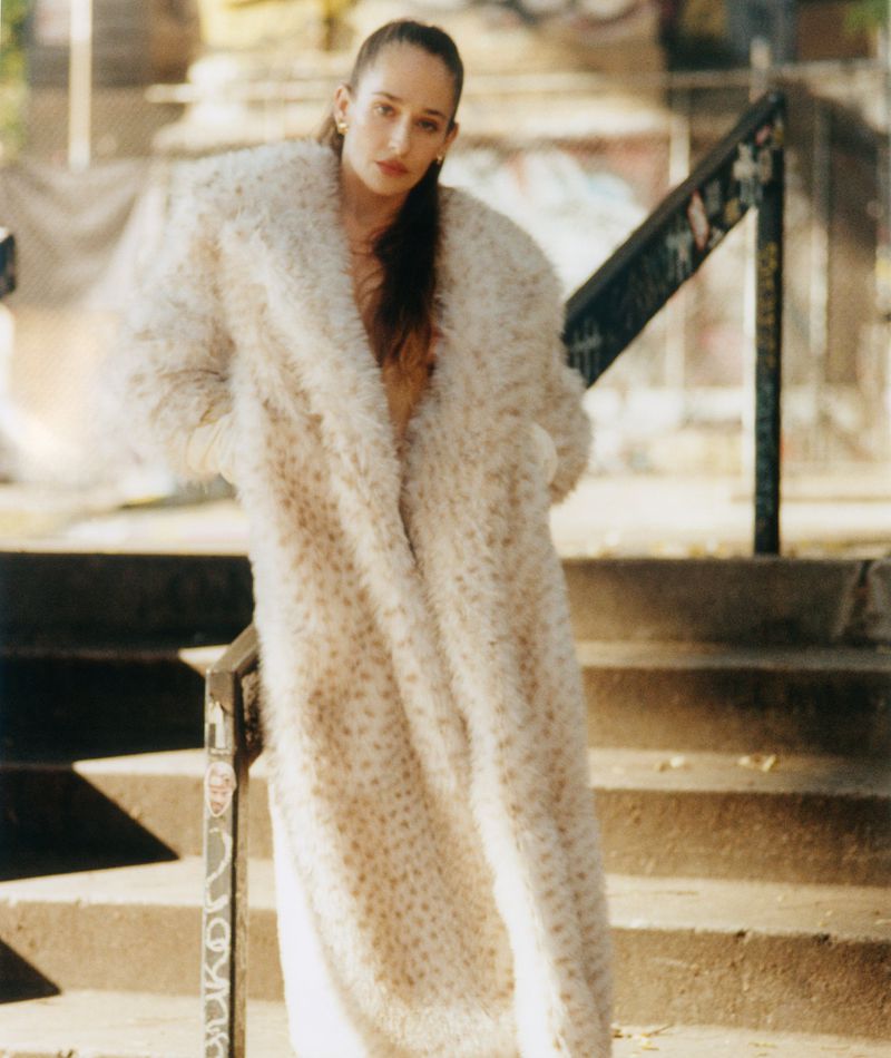 Jemima Kirke wearing a long cream faux-fur coat posing on outdoor urban stone steps for an editorial portrait.