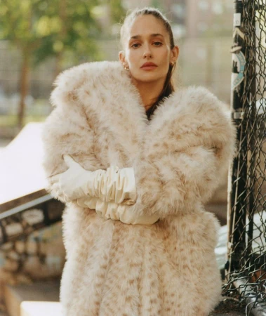 Jemima Kirke wearing a fluffy textured coat and white gloves standing outdoors near a chain-link fence.