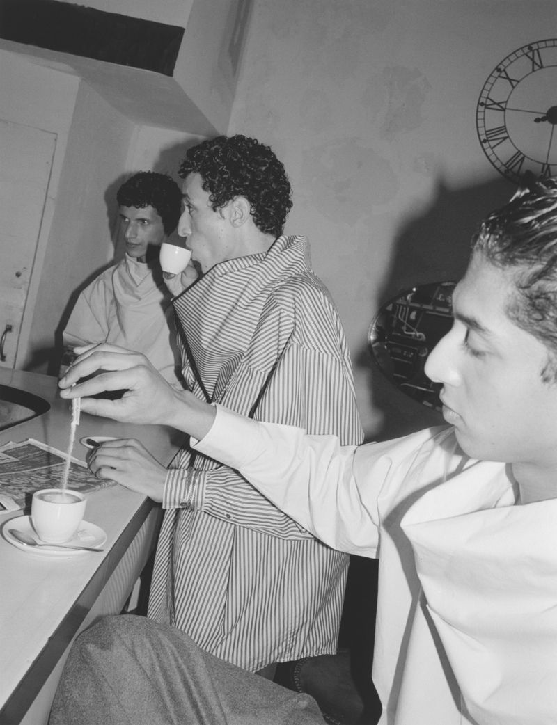 Black and white indoor shot of three adult males at a bar, one drinking from a coffee cup with a straw, featuring a vintage aesthetic.