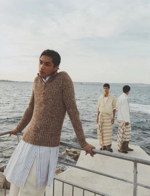 Three men in stylish casualwear pose on a concrete platform near the ocean on a cloudy day, showcasing relaxed yet fashionable attire.