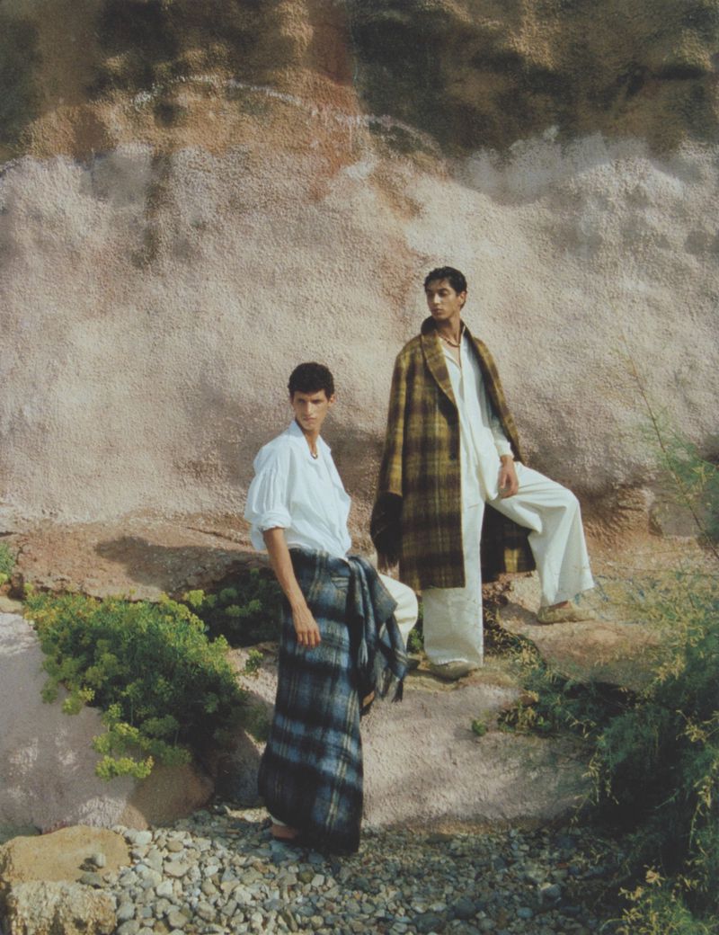 Two adult males in stylish clothing pose outdoors against a textured wall with natural greenery. Fashion editorial, lifestyle theme.