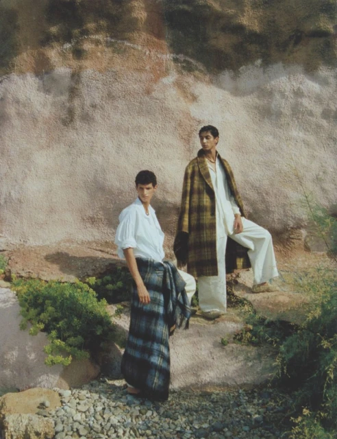 Two adult males in stylish clothing pose outdoors against a textured wall with natural greenery. Fashion editorial, lifestyle theme.
