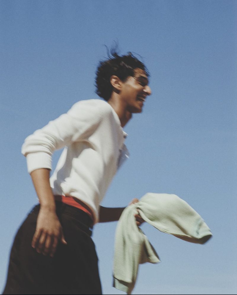 Smiling adult male holding light green towel against a clear blue sky, wearing a white long-sleeve shirt and dark pants in outdoor setting.
