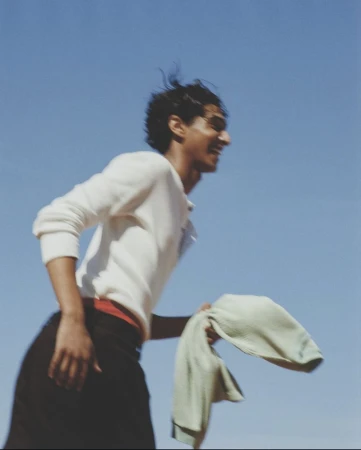 Smiling adult male holding light green towel against a clear blue sky, wearing a white long-sleeve shirt and dark pants in outdoor setting.