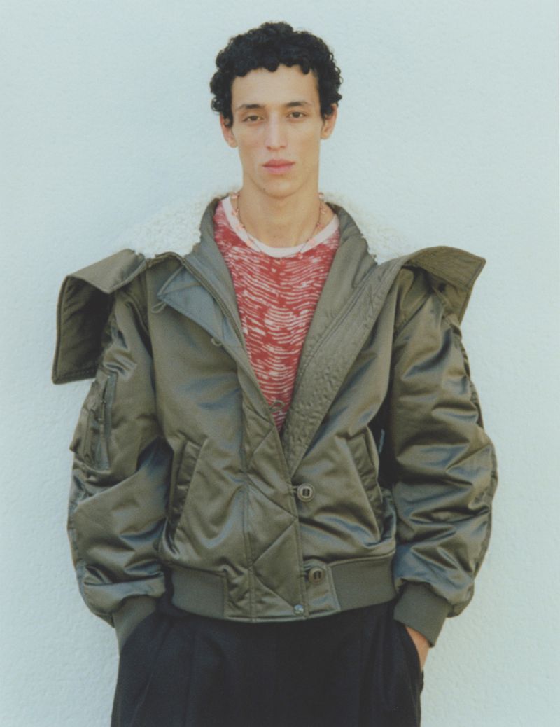 A young adult male model poses in an olive green bomber jacket and black pants against a pale neutral background in a full shot.