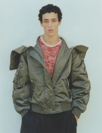 A young adult male model poses in an olive green bomber jacket and black pants against a pale neutral background in a full shot.