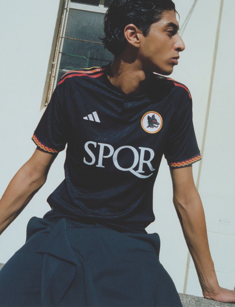 Young adult male model wears an Adidas Roma soccer jersey, seated against a white wall in casual outdoor fashion shot.