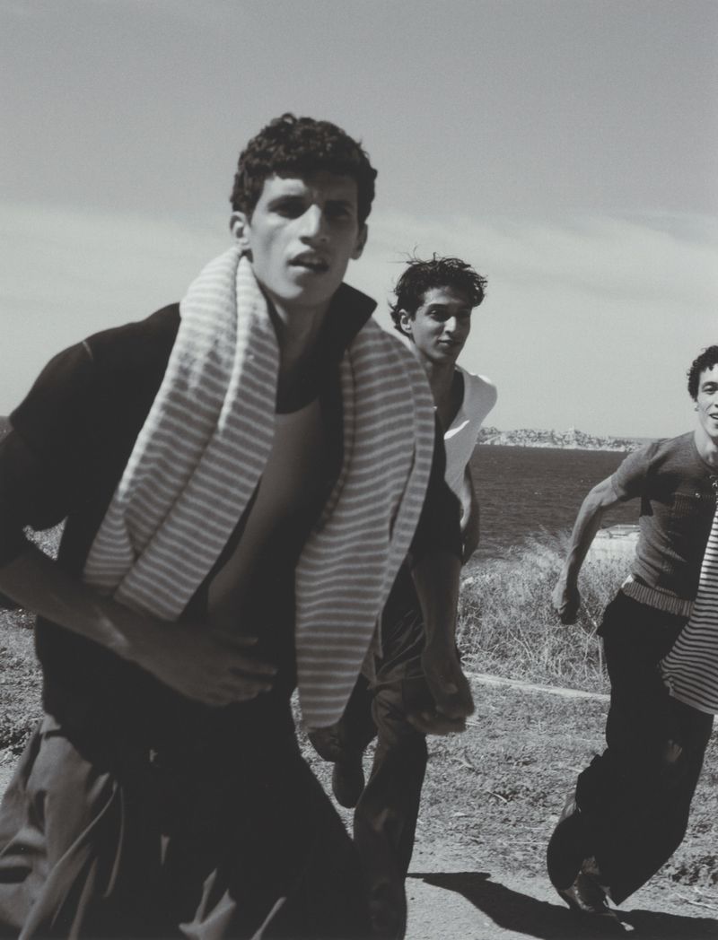 Three men in casual attire run outdoors on a grassy path. A striped towel is draped around one man's neck on a sunny day, monochrome.