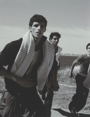 Three men in casual attire run outdoors on a grassy path. A striped towel is draped around one man's neck on a sunny day, monochrome.