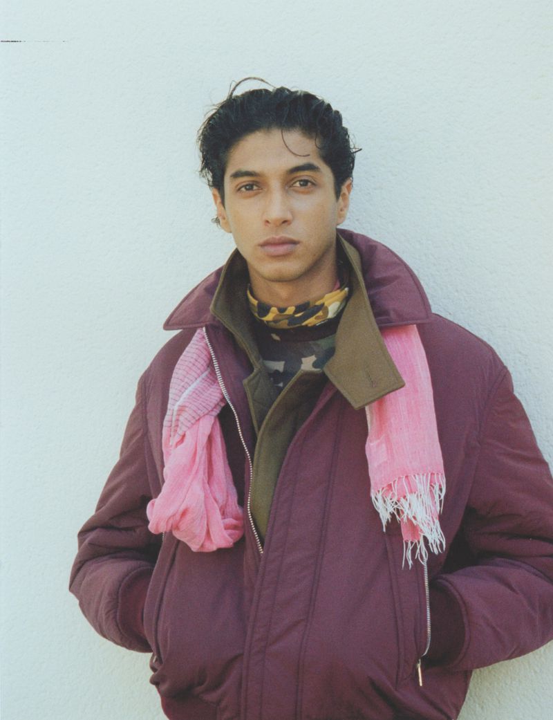 A young adult male stands indoors wearing a maroon jacket and a colorful scarf, posing in front of a white wall in a medium shot.