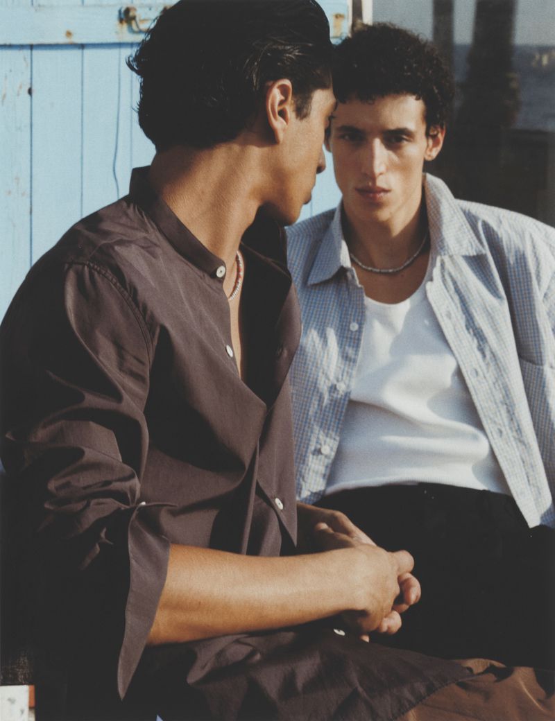 Two adult males in casual summer clothing, outdoors with natural light, standing close together, on blue wall background.