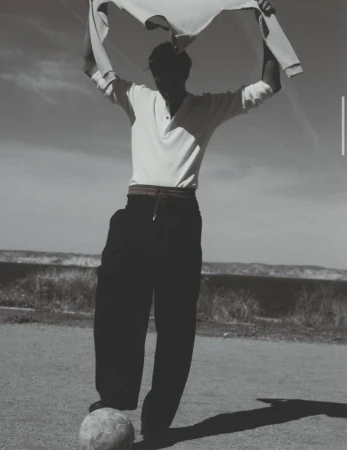 Monochrome fashion editorial image of a male model holding a sweater above his head and resting his foot on a soccer ball outdoors.
