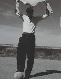Monochrome fashion editorial image of a male model holding a sweater above his head and resting his foot on a soccer ball outdoors.