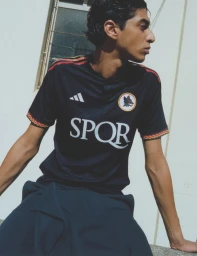 Young man in a black SPQR soccer jersey seated against a sunlit exterior building wall and window.