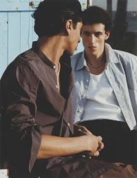 Two young men in casual shirts seated outdoors against a pale blue wooden wall in a fashion editorial portrait.