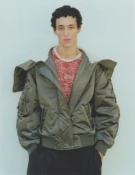A male model wearing an oversized olive green bomber jacket with a shearling collar against a plain light background.