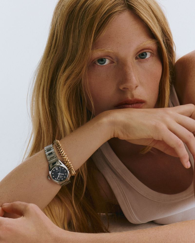 Close-up of a fair-skinned, red-haired adult female posing with a luxury watch and gold bracelet against a bright white backdrop.