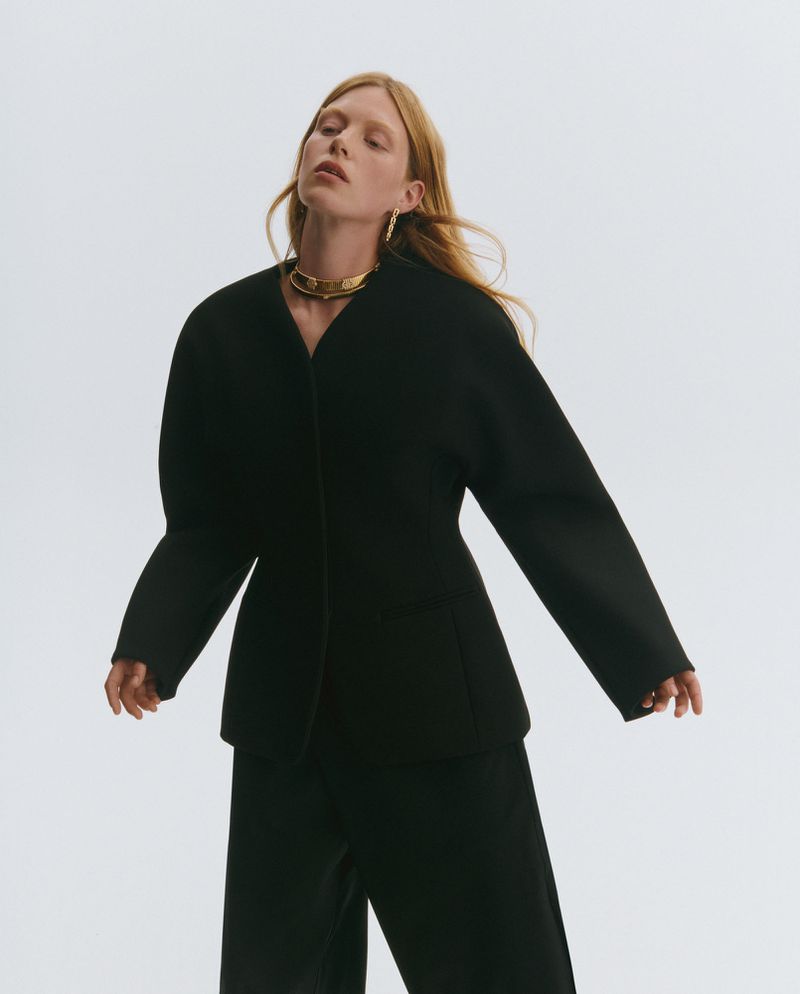 Fashion shot of a woman in a black suit with a gold choker and light hair posing in a studio setting with a white background.
