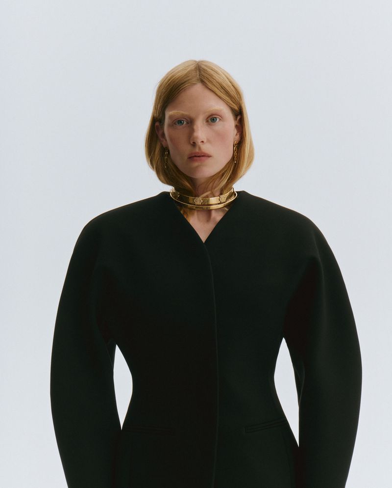 A fair-skinned woman with auburn hair poses indoors wearing a black jacket and gold necklace against a solid white backdrop.