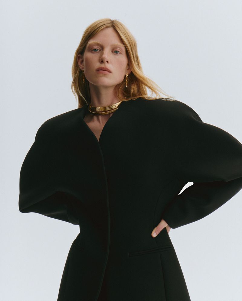 Woman with fair skin and blonde hair poses wearing a black top and gold choker in a studio setting with a white background.