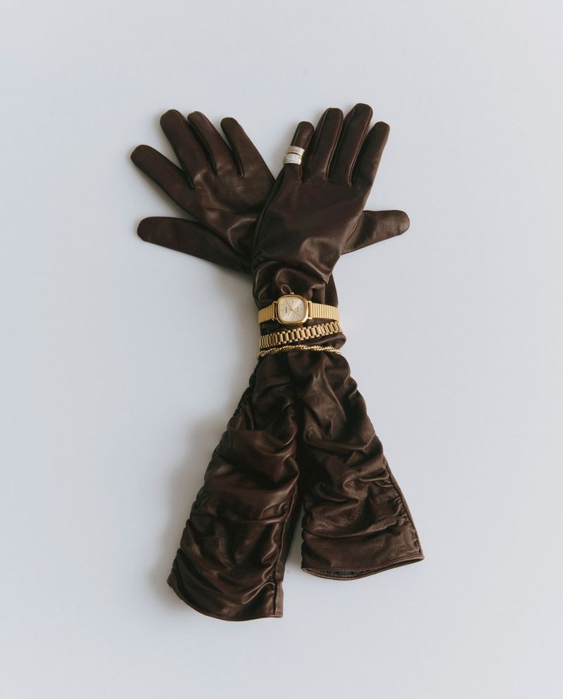 Long brown ruched leather gloves styled with a gold wristwatch, chain bracelets, and a ring on a plain white surface.