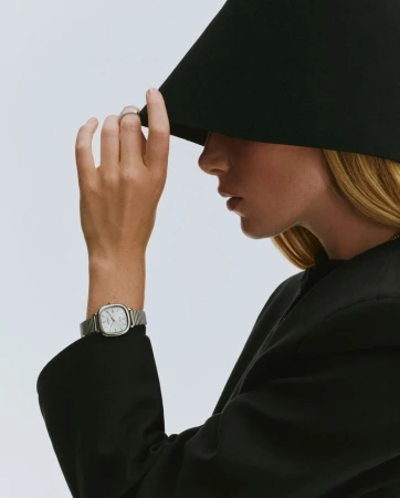 A woman in profile wearing an oversized black cone hat, black blazer, and silver wristwatch against a clean light background.