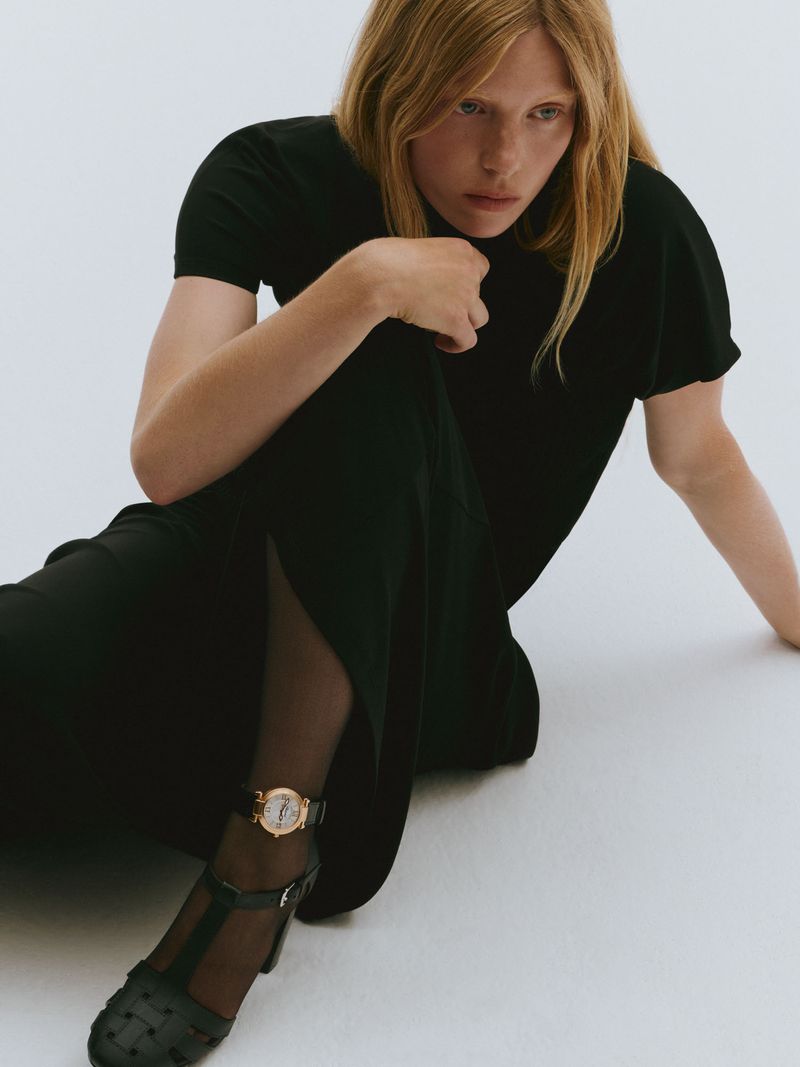 Woman in black dress and tights sitting on white background, wearing a gold watch and black heels, in a fashion studio shot.