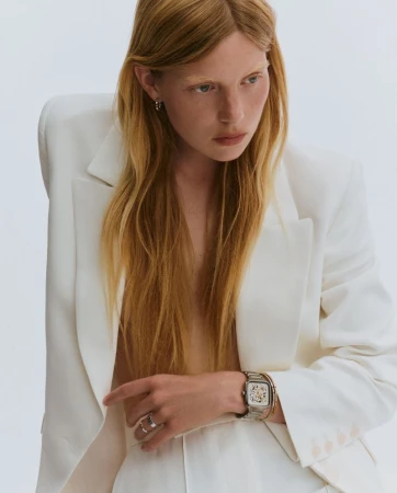 Blonde woman in white pantsuit, showcasing a wristwatch. Studio lighting and simple background highlight the fashion and accessory details.