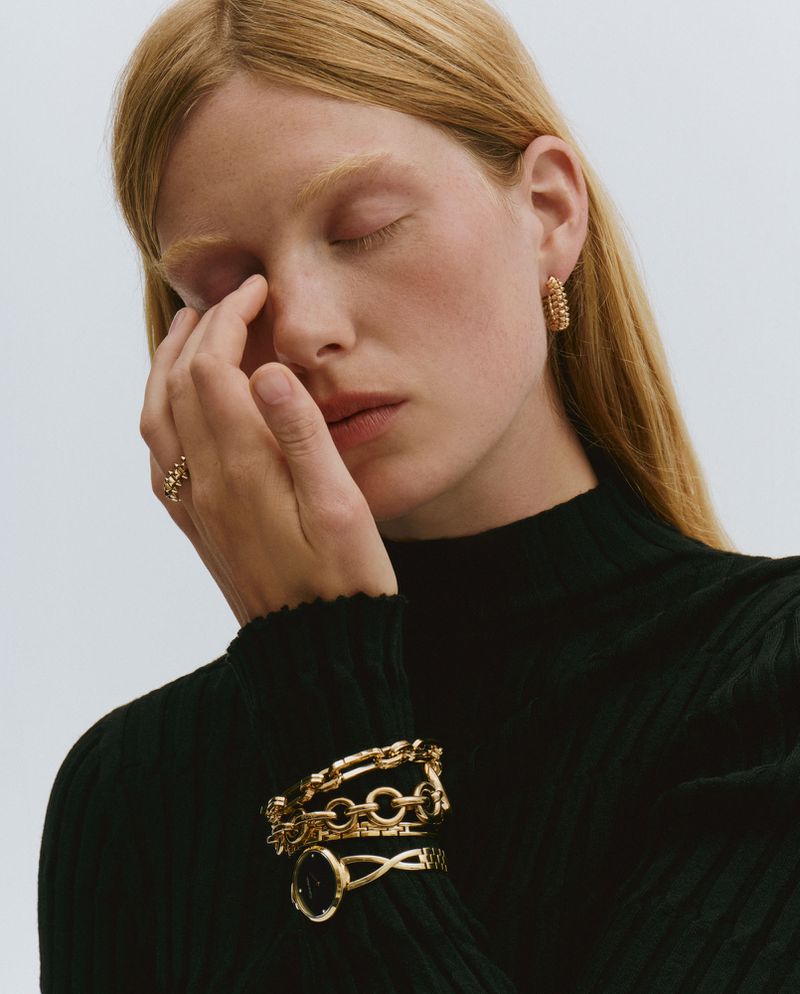 Portrait of a blond woman wearing gold earrings, ring, and bracelets, with her eyes closed and her hand near her face in a studio setting.