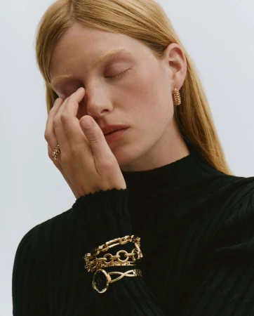 Portrait of a blond woman wearing gold earrings, ring, and bracelets, with her eyes closed and her hand near her face in a studio setting.