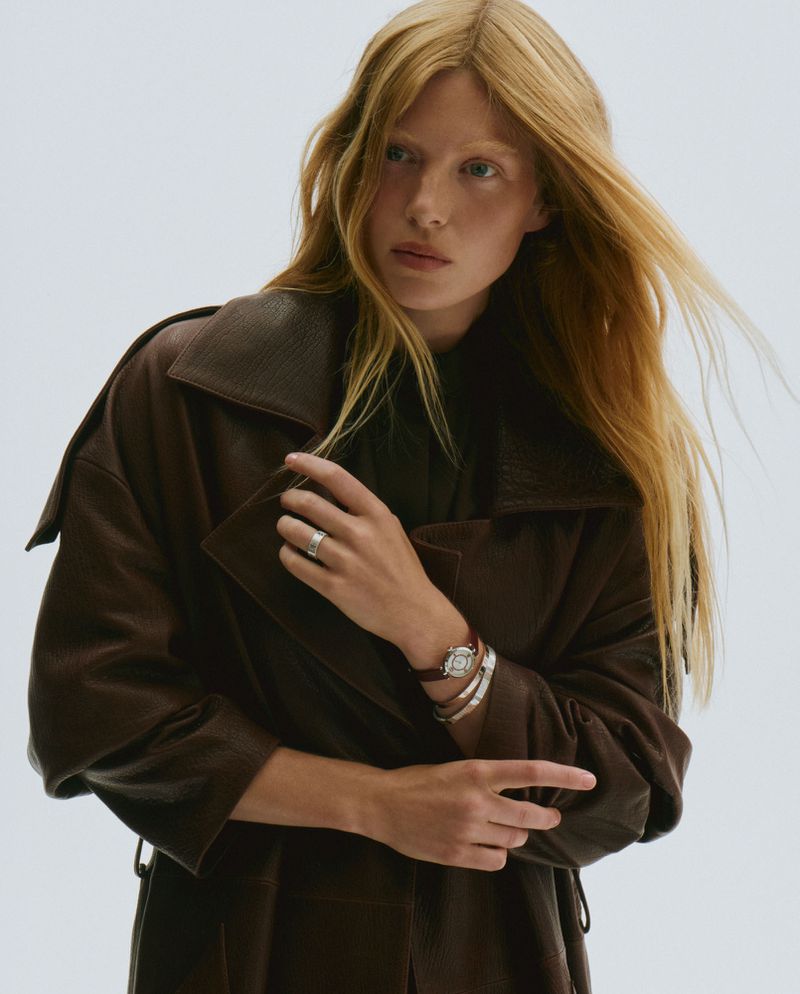 Portrait of a fair-skinned female model with long blonde hair wearing a brown leather trench coat against a bright white background.