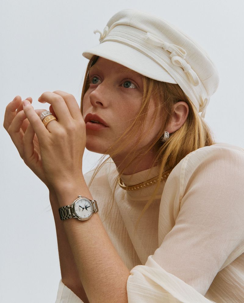 Portrait of a young woman with a white hat, jewelry, and a watch, looking upward against a light gray background in a fashion editorial.