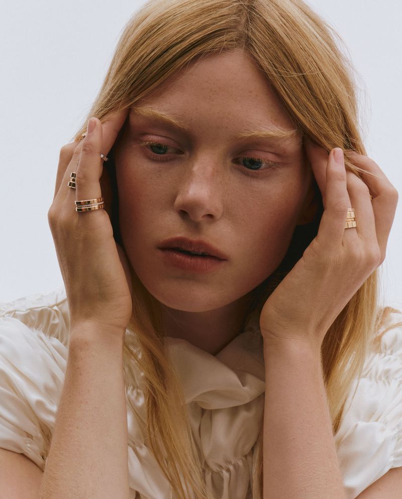 Close-up portrait of a blonde woman with fair skin wearing gold rings and a white blouse, set against a light background.