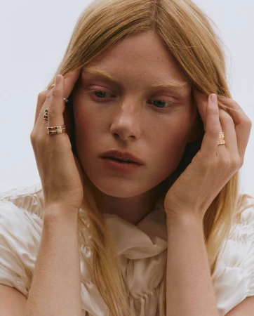 Close-up portrait of a blonde woman with fair skin wearing gold rings and a white blouse, set against a light background.