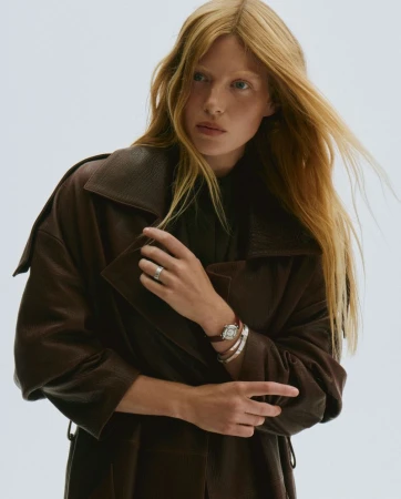 Blonde model wearing a dark brown oversized leather coat, silver ring, and luxury wrist watch against a pale seamless studio background.