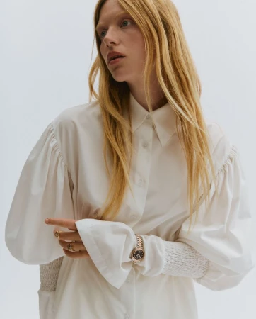 Model with long blonde hair wearing a white collared blouse, gold rings, and a metal watch against a white background.