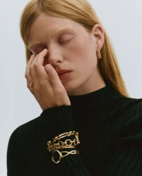 Model Tes Linnenkoper wears a black turtleneck, stacked gold chain bracelets, watch, and hoop earrings against a neutral backdrop.