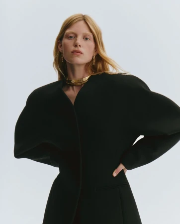 Model Tes Linnenkoper in a black sculptural coat with wide shoulders and a gold choker necklace against a white background.