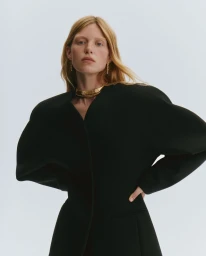 Model Tes Linnenkoper in a black sculptural coat with wide shoulders and a gold choker necklace against a white background.