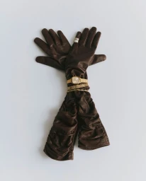 Long brown ruched leather gloves styled with a gold wristwatch, chain bracelets, and a ring on a plain white surface.