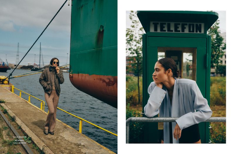 Editorial fashion shoot on a pier with a model in a jacket by a large ship; a woman in a phone booth leans on railings.