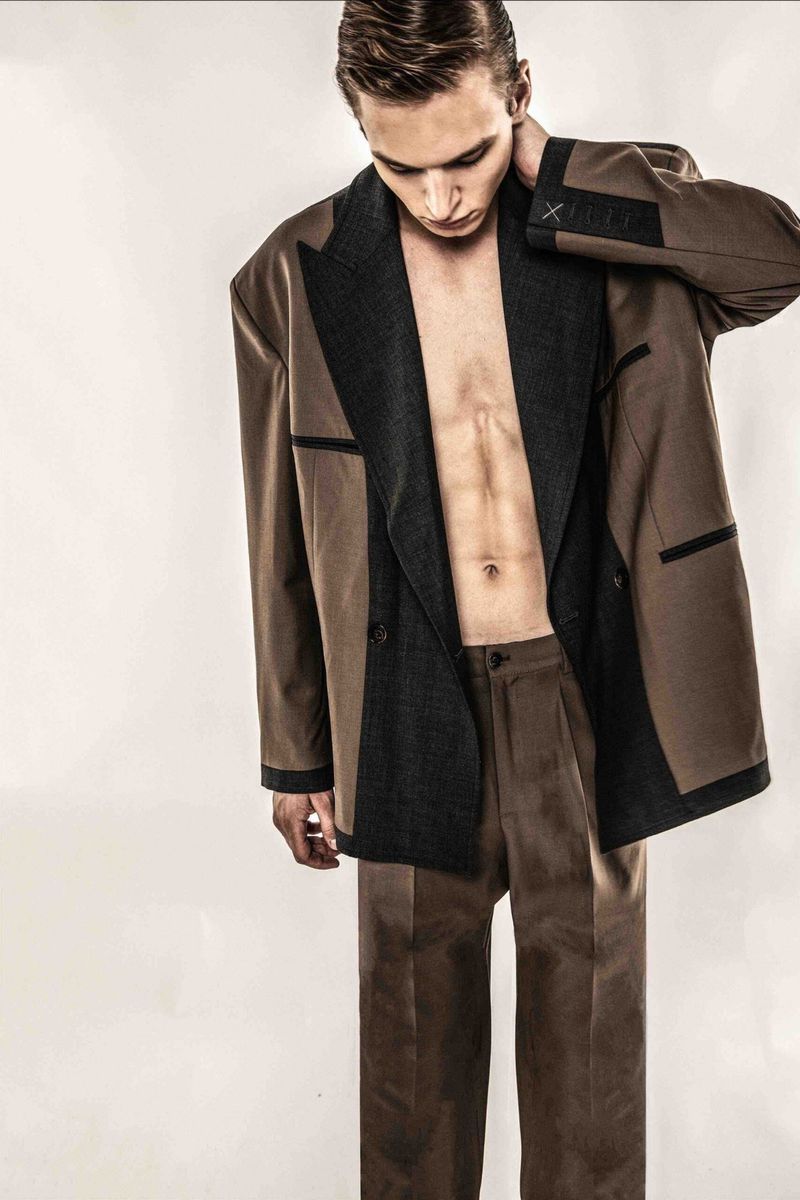 A young adult male poses with an open brown and gray suit jacket, against a plain white backdrop.