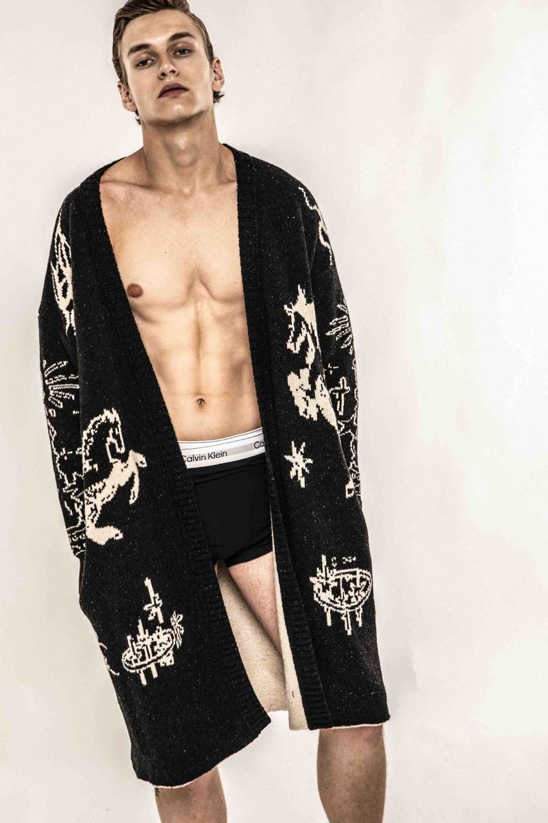 Male model poses indoors wearing a patterned black cardigan and Calvin Klein branded underwear, showcasing fashion and physique.