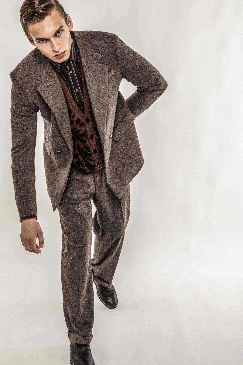 A young male fashion model poses in a tweed suit, brown patterned vest, striped shirt and black shoes against a white backdrop.