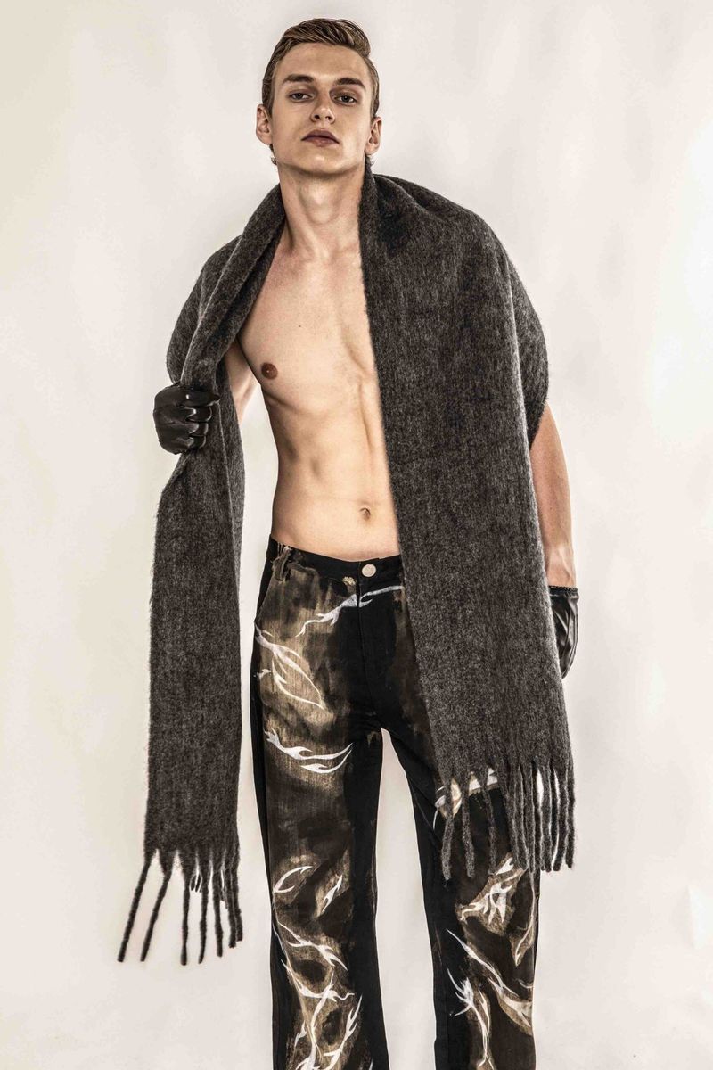 A slender male fashion model poses shirtless in a photo studio wearing a grey fringe scarf and black patterned pants against a white backdrop.