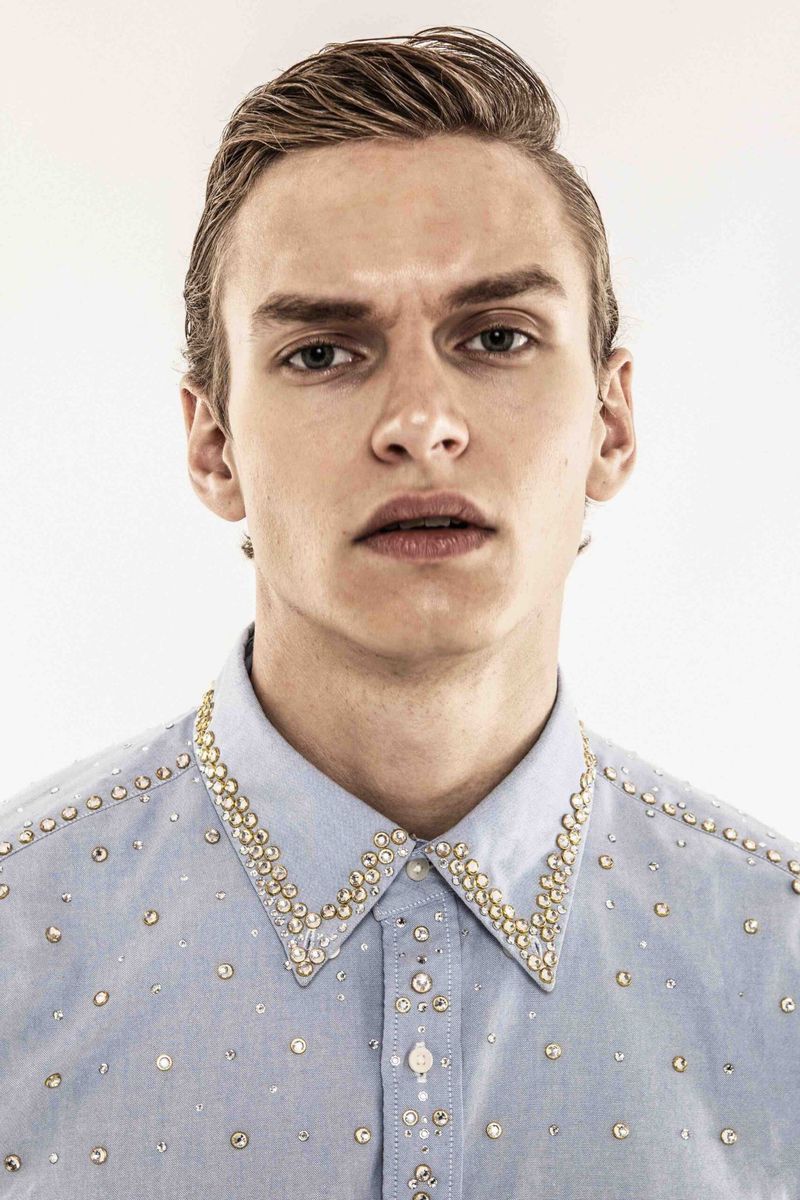 Close-up of a young adult male model with blond hair wearing a light blue button-down shirt with rhinestones on a white background.