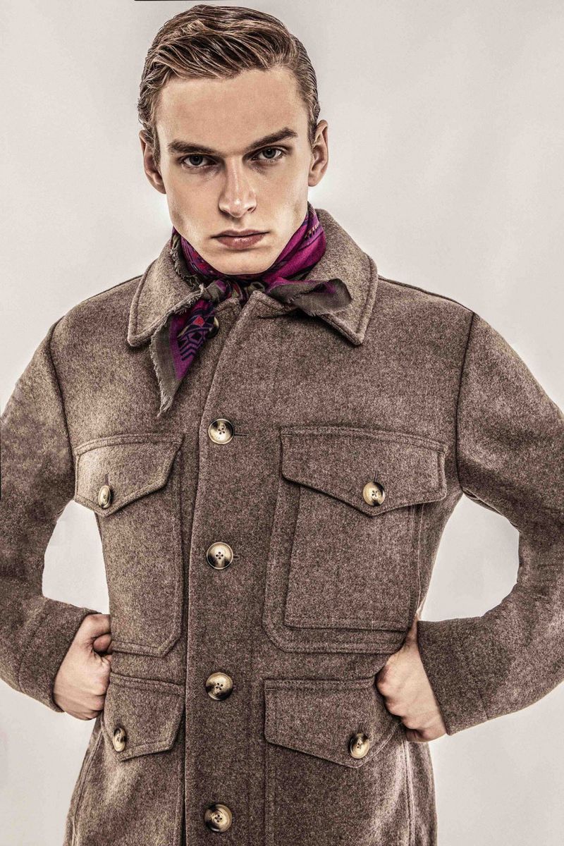 Portrait of a stylish man in a brown wool coat and purple scarf, posing indoors against a pale background in a fashion shot.