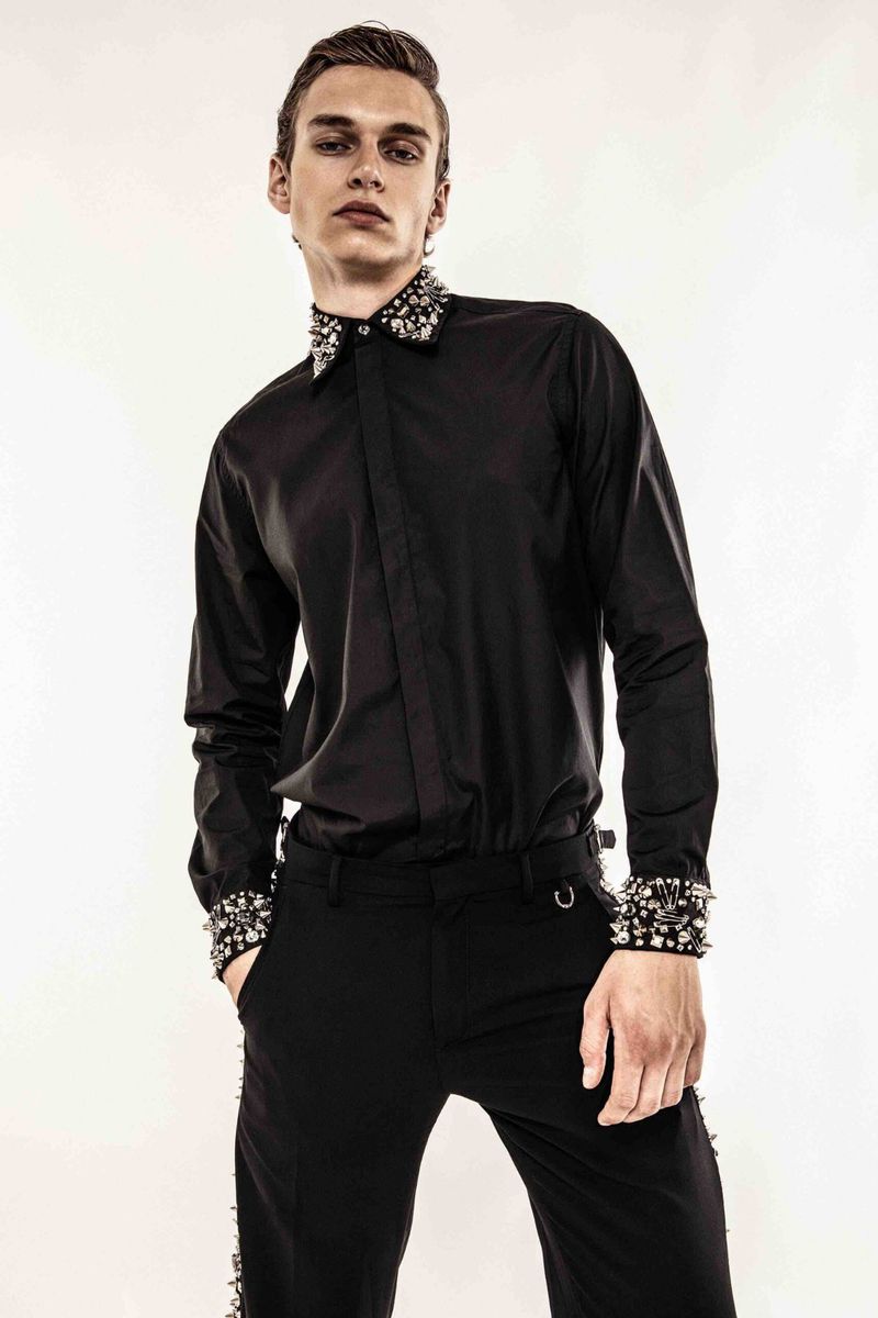 A stylish male fashion model poses in a black button-down shirt with studded collar and cuffs, paired with black pants on a white backdrop.