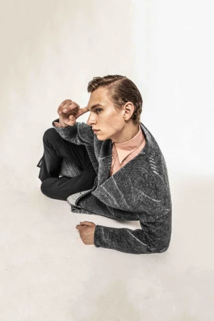 Model Jan Rykowski reclining in a textured grey wrap jacket and peach collared shirt against a white studio floor.