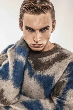 Close-up portrait of a young male model wearing a textured blue and brown patterned sweater against a neutral background.